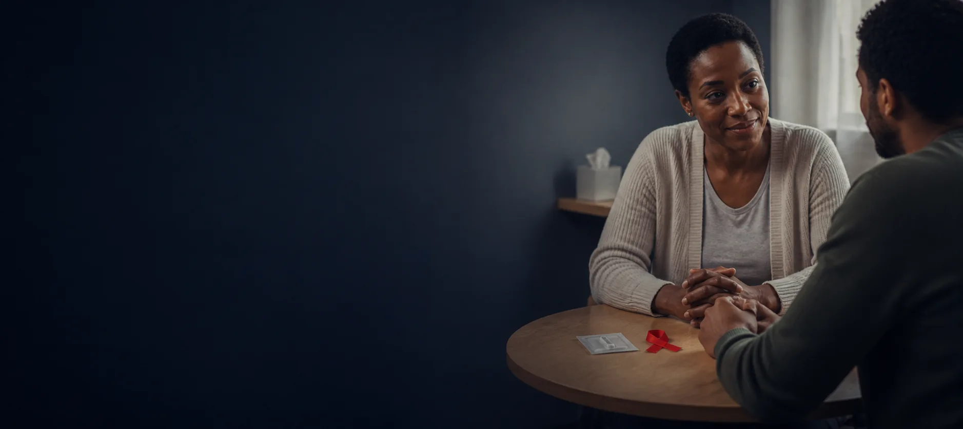 A PHC clinician sitting with a patient, hands held, a red ribbon on the table